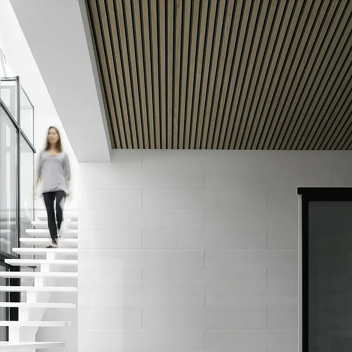 Grosfillex Wandpaneel Element 3D - Linio Walnut, 2.600 x 375 x 8 mm - Innenaufnahme mit einer Frau, die eine Treppe in einem modernen Haus mit Holzdecke und weißem Wandfliesendesign hinuntergeht.