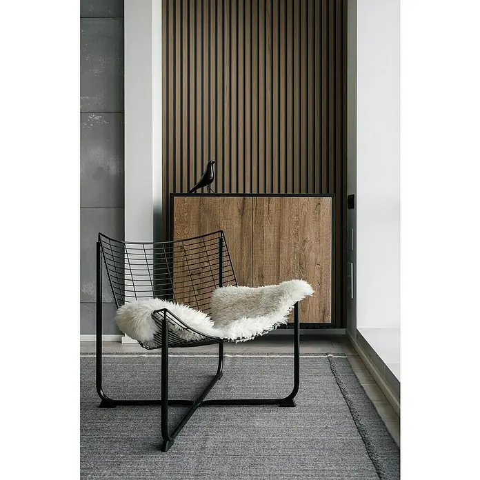 Grosfillex Wandpaneel Element 3D - Linio Walnut, 2.600 x 375 x 8 mm - Stilvolles Interieur mit einem schwarzen Drahtsessel und einem weichen Fellkissen auf einem grauen Teppich vor einer Holzvertäfelung.
