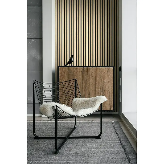 Grosfillex Wandpaneel Element 3D - Linio Ash, 2.600 x 375 x 8 mm - Das Bild zeigt einen modernen schwarzen Drahtsessel mit einem flauschigen Kissen auf einem grauen Teppich, vor einer Holzvertäfelung und einem Schrank.