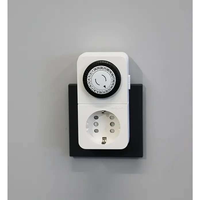 UniTEC Tages-Zeitschaltuhr Greenline Analog - Weiß, 3.680 W - Nahaufnahme einer Zeitschaltuhr mit einer schwarzen Blende und einer weißen Steckdose auf einer grauen Wand.