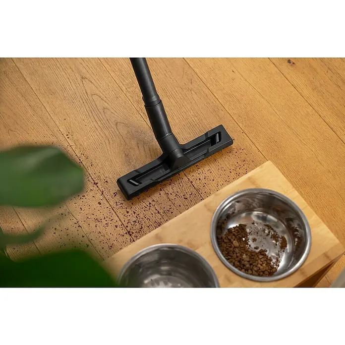Kärcher Waschsauger SE 4 GO!Further - 1.000 W, Behältervolumen: 4 l - Ein Staubsauger reinigt den Boden neben zwei Hundenäpfen, die auf einem Holzbrett stehen. Einige Krümel liegen um die Basis des Staubsaugers.
