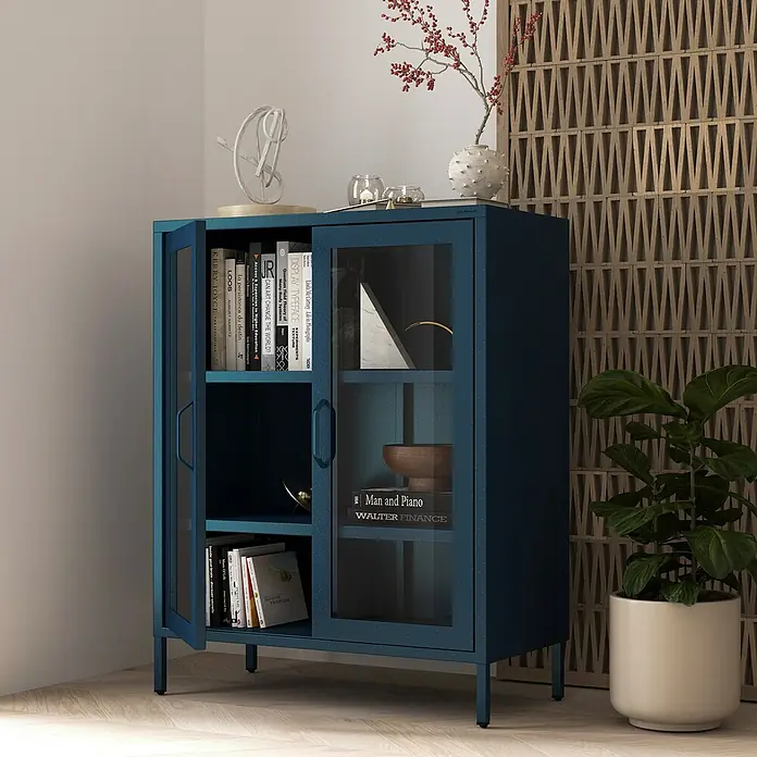 Jan Nowak Vitrine Gina - B x T x H: 80 x 40 x 101,5 cm, Dunkelblau - Blauer Metallschrank mit Glastüren, gefüllt mit Büchern und dekorativen Gegenständen, neben einer Zimmerpflanze.
