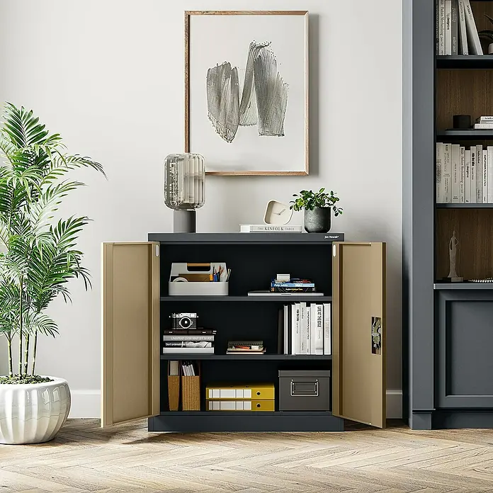 Jan Nowak Aktenschrank Beata - B x T x H: 90 x 40 x 92,5 cm, Anthrazit / Nussbaum - Ein geräumiger Büroschrank in Anthrazit mit offenen beigefarbenen Türen, gefüllt mit Büchern und Deko, unter einem modernen Kunstwerk.