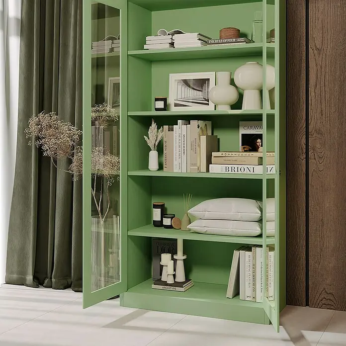 Jan Nowak Aktenschrank Amelia - B x T x H: 90 x 40 x 185 cm, Grün - Ein grünes Bücherregal mit Glasfront, gefüllt mit Büchern, Vasen und Dekorationsgegenständen, steht vor einer grünen Vorhangfassade.