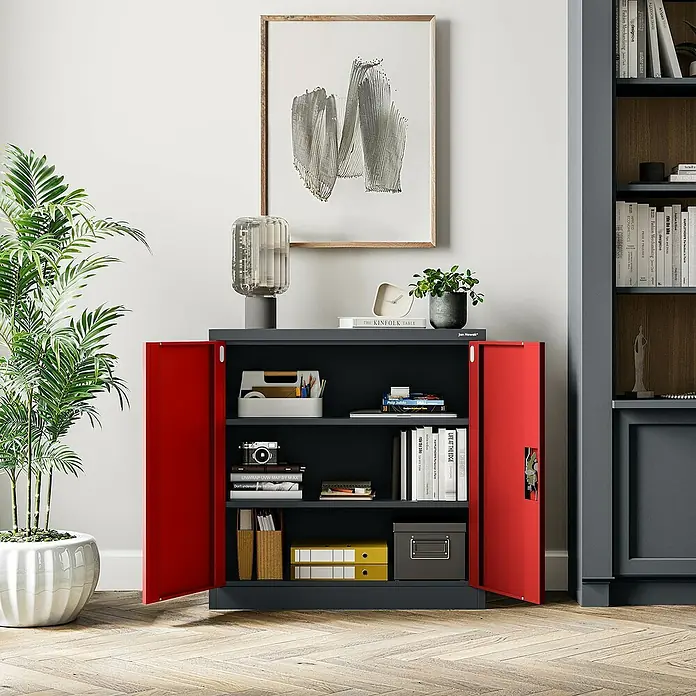 Jan Nowak Aktenschrank Beata - B x T x H: 90 x 40 x 92,5 cm, Anthrazit / Rot - Ein grauer Aktenschrank mit roten Türen ist mit Büchern und Dekorationsgegenständen gefüllt. Ein Kunstwerk hängt darüber.