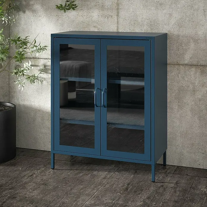 Jan Nowak Vitrine Gina - B x T x H: 80 x 40 x 101,5 cm, Dunkelblau - Blauer Glasschrank mit zwei Türen auf einem strukturierten Untergrund, neben einer grünen Pflanze.