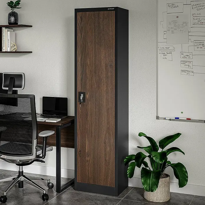 Jan Nowak Aktenschrank Alex - B x T x H: 45 x 40 x 185 cm, Anthrazit/ Nussbaum - Ein schlanker, brauner Holzschrank mit schwarzem Rahmen steht in einem hellen Raum, neben einem Schreibtisch und einer Büropflanze.