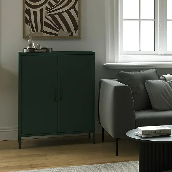 Jan Nowak Kommode Vito - B x T x H: 80 x 40 x 101,5 cm, Flaschengrün - Ein dunkelgrüner Metallschrank mit zwei Türen steht neben einem grauen Sofa und einem runden Tisch. Dekorative Elemente sind auf dem Schrank platziert.
