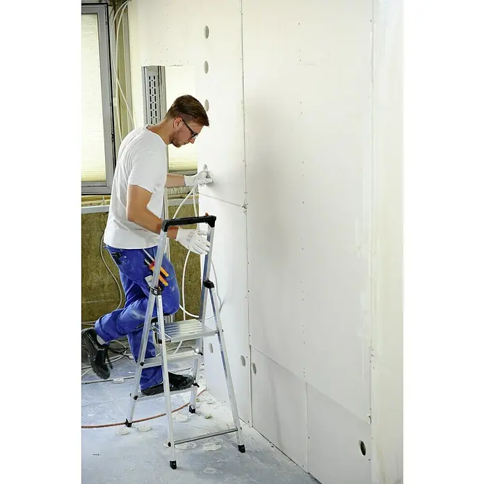 Krause Monto Stehleiter Solidy - Arbeitshöhe: 2,65 m, Anzahl Tritte: 3 Stk., Belastbarkeit: 150 kg, Aluminium - Mann auf einer Leiter, der an einer Wand mit Löchern arbeitet. Deutlich ist die Baustellensituation mit sichtbarer Dämmung und Elektrik.