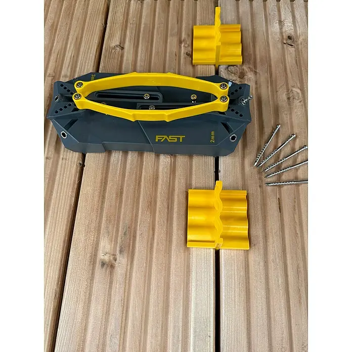 Fast Bohrhilfe - L x B x H: 190 x 90 x 70 mm, Grau/Gelb - Das Bild zeigt ein graues Terrassendielen-Befestigungswerkzeug mit gelben Akzenten, zwei gelbe Abstandshalter und vier Schrauben auf einem Holzboden.
