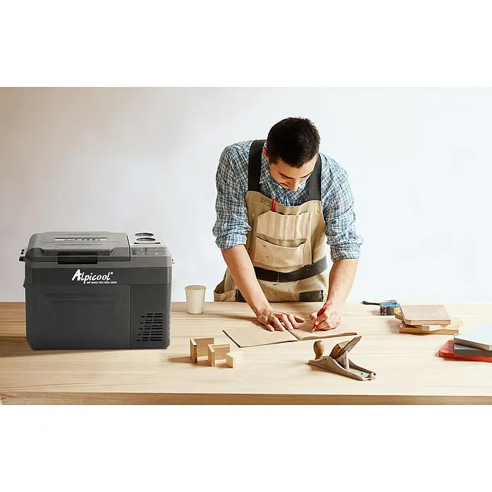 Kompressor Kühlbox WTW25 - 593 x 345 x 410 mm - Ein Alpicool Kühlbox neben einem Handwerker, der an einem Tisch mit Werkzeugen arbeitet. Ideal für unterwegs, hält Speisen und Getränke kühl.