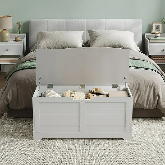 Truhe - L x B x H: 40 x 100 x 46 cm, Weiß - Eine offene, weiße Aufbewahrungsbox aus Holz vor einem Bett. Die Truhe enthält Kleidung und ist ideal zur Aufbewahrung im Schlafzimmer.