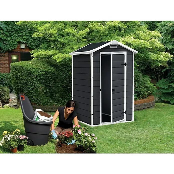 Keter Gerätehaus Manor 4x3 - Außenmaß inkl. Dachüberstand (B x T): 129 x 103 cm, Kunststoff, Anthrazit/Weiß - Eine Frau gärtnert vor einem anthrazitfarbenen Gartenschuppen mit weißen Akzenten, der im Garten steht.