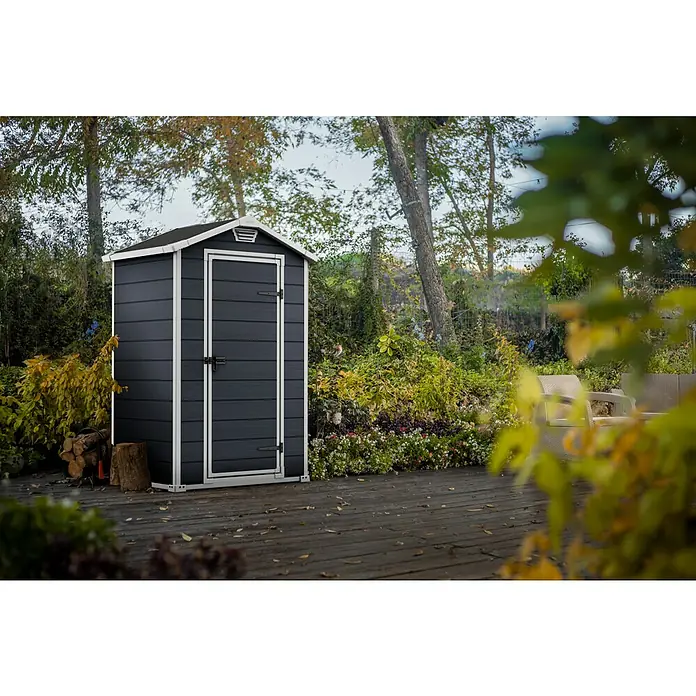 Keter Gerätehaus Manor 4x3 - Außenmaß inkl. Dachüberstand (B x T): 129 x 103 cm, Kunststoff, Anthrazit/Weiß - Das Bild zeigt ein graues Gartenhaus mit weißen Akzenten auf einer hölzernen Terrasse, umgeben von üppigem Grün und Bäumen.