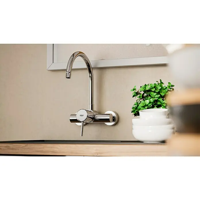 Grohe Concetto Spültischarmatur - Chrom, Glänzend, Wandmontage - Nahaufnahme einer glänzenden Grohe Küchenarmatur an einer Wand mit Waschbecken und dekorativen Schüsseln sowie einer Pflanze.
