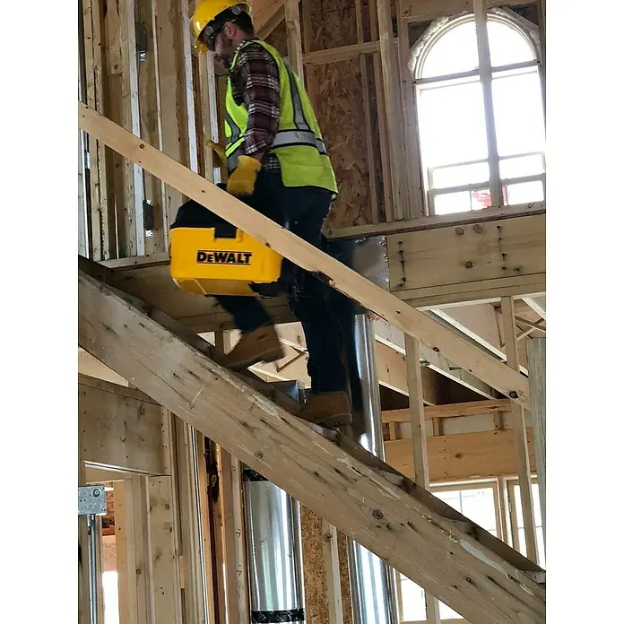DeWalt Elektro-Nass- & Trockensauger DXV23G - 230 W, Behältervolumen: 23 l - Ein Bauarbeiter mit einem gelben DeWalt Werkzeugkasten steigt eine Treppe auf einer Baustelle hinauf, Schutzhelm und Warnweste inklusive.