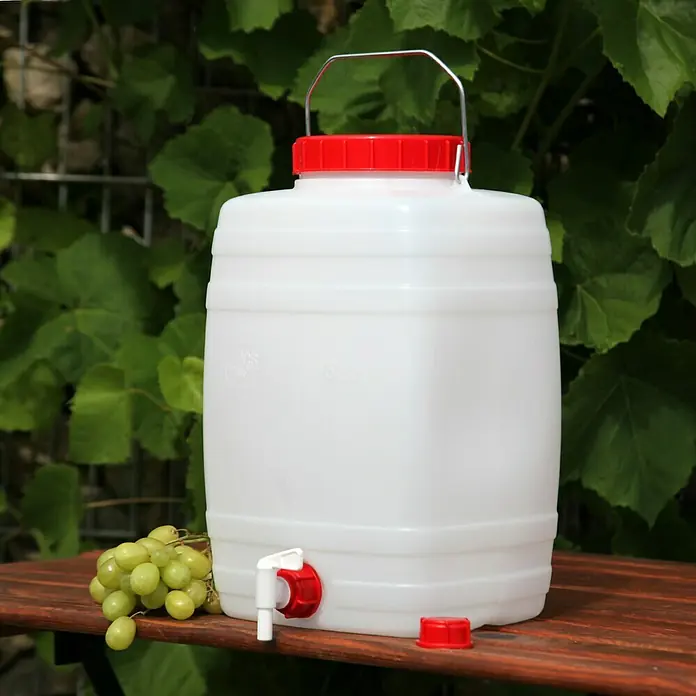 Floraworld Getränkefass Standard - Fassungsvermögen: 15 l - Ein weißer Kunststoffbehälter mit rotem Deckel und Hahn, ideal für Flüssigkeiten, steht auf einem Holztisch neben einer Traube.