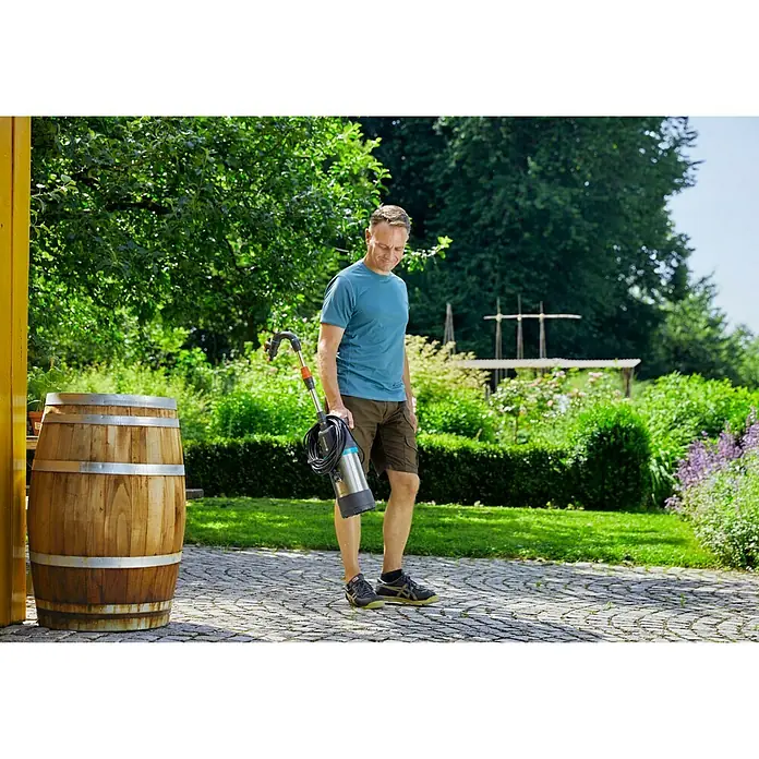 Gardena Regenfasspumpe 4700/2 inox automatic - Leistung: 550 W, Max. Fördermenge: 4.700 l/h - Ein Mann trägt eine GARDENA Regenfasspumpe in einem Garten, neben einem Holzfass. Perfekt für die Gartenbewässerung.