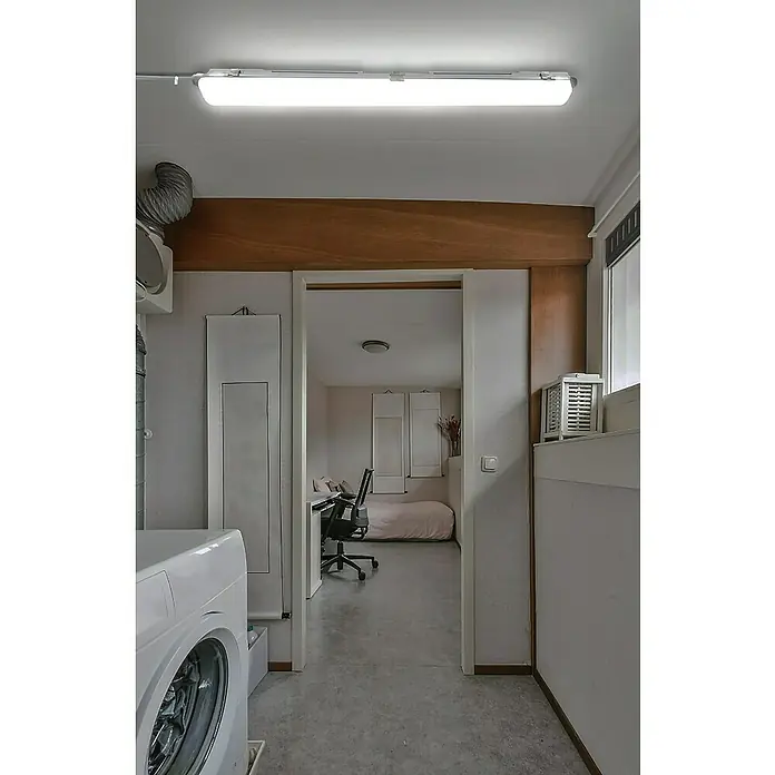 Voltolux Feuchtraumleuchte - L x B x H: 128,4 x 6,2 x 6 cm, Lichtfarbe: Tageslichtweiß, 14 W, IP65 - Blick durch einen Durchgang, der eine Waschmaschine, einen Schreibtisch und ein Bett zeigt. Beleuchtet wird der Raum durch eine Neonröhre.