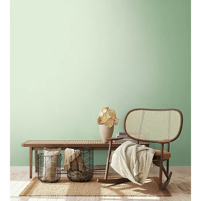 A.S. Création Aquarell Dreams Vliestapete - Hellgrün, Uni, 10,05 x 0,53 m - Elegantes Interieur mit Rattanschaukelstuhl, Holzbank und dekorativen Körben auf einem Jute-Teppich vor einer mintgrünen Wand.