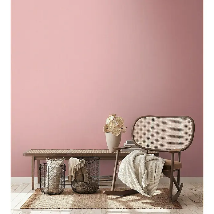 A.S. Création Aquarell Dreams Vliestapete - Rosa, Uni, 10,05 x 0,53 m - Gemütliche Einrichtung mit einem Rattansessel, einem Holzbänkchen, Körben und einer Vase auf einem Teppich vor rosa Wand.