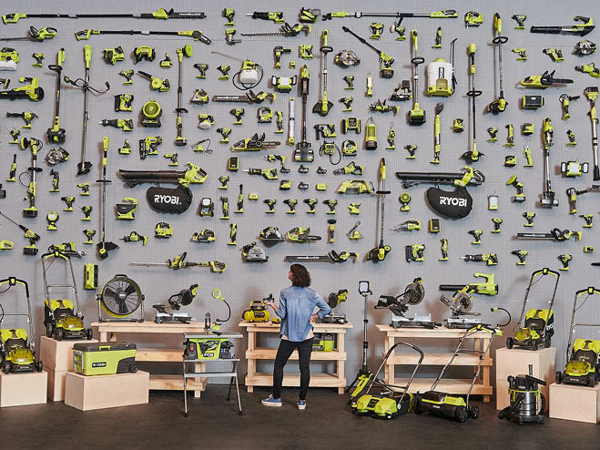 Frau steht vor einer grauen Wand, die mit einer großen Auswahl an Ryobi Produkten behangen ist. Auf kleinen Kommoden stehen Rasenmäher
