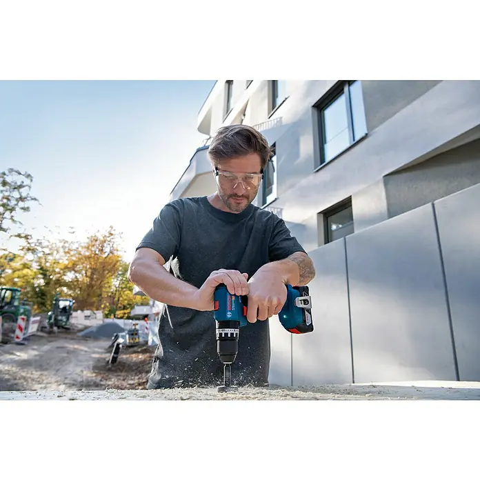 Bosch Professional AMPShare 18V Akku-Schlagbohrschrauber GSB 18V-45 - 18 V, 1 Akku, 2 Ah - Ein Mann bohrt mit einem blauen Bosch Akku-Bohrschrauber in ein Holzbrett, während er eine Schutzbrille trägt.