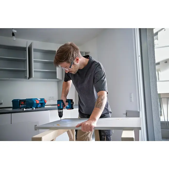 Bosch Professional AMPShare 18V Akku-Schlagbohrschrauber GSB 18V-45 - 18 V, 1 Akku, 2 Ah - Ein Handwerker bohrt mit einem blauen Bosch-Akkuschrauber ein Loch in eine weiße Platte. Er trägt eine Schutzbrille und hat eine Werkzeugkiste im Hintergrund.