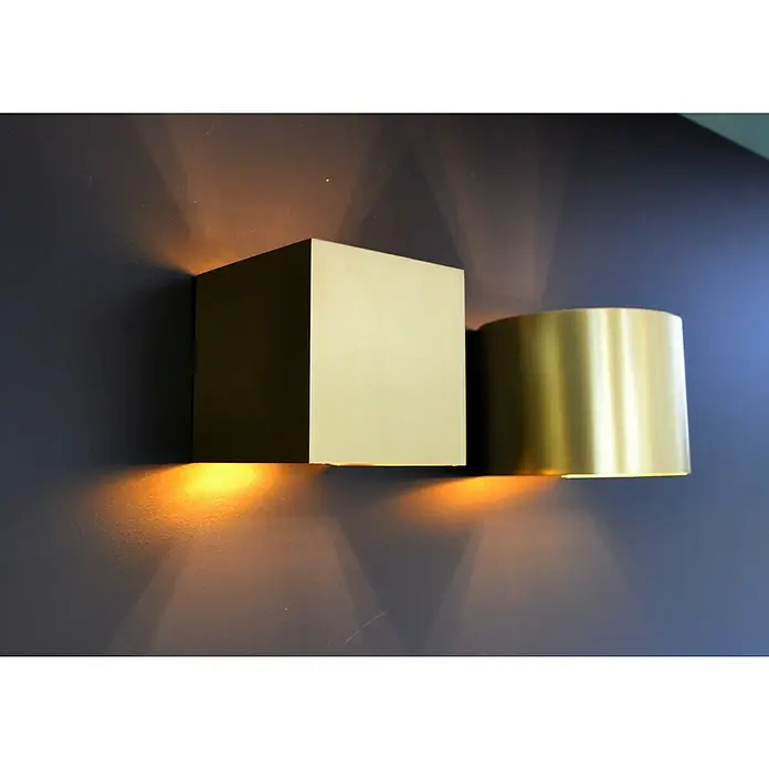 Lucide LED-Wandleuchte Xio - 3,5 W, L x B x H: 14 cm x 12 cm x 10,5 mm, Gold, Warmweiß - Eine moderne Wandleuchte in Goldoptik, bestehend aus einem Würfel und einem Zylinder, die warmes Licht an eine blaue Wand wirft.