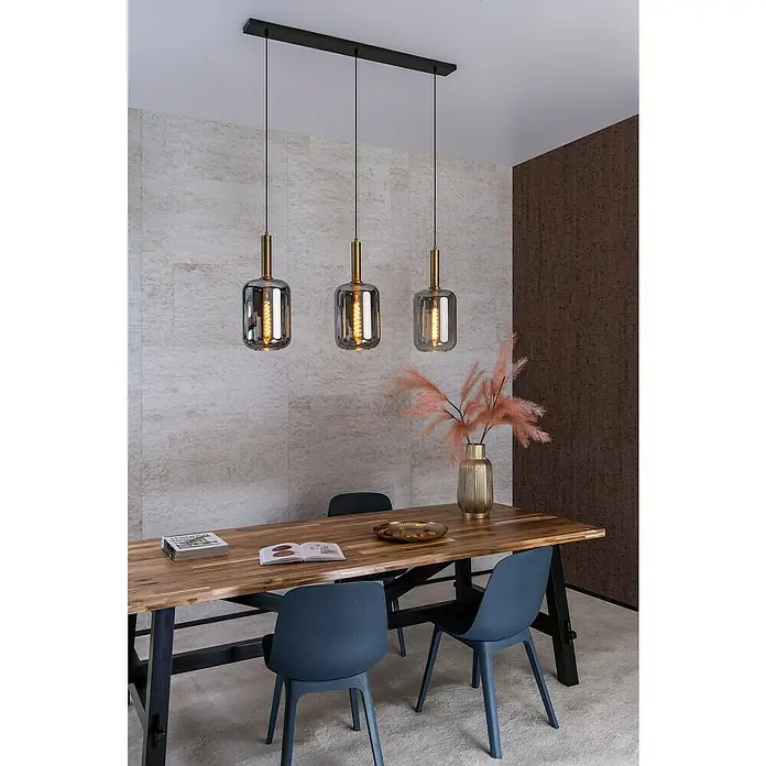 Lucide Pendelleuchte Joanet - 40 W, L x B x H: 110 x 22 x 150 cm, Rauchfarben, E27, Anzahl Leuchtmittel: 3 Stk. - Dreifach-Pendelleuchte mit grauen Glasschirmen und goldfarbenen Akzenten über einem Holztisch mit blauen Stühlen.