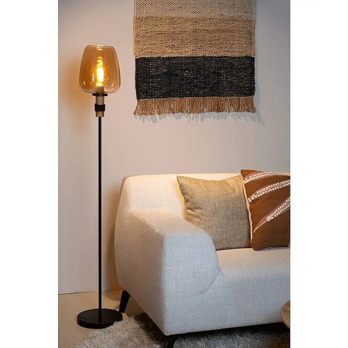 Lucide Ilona Stehleuchte - 40 W, Höhe: 150 cm, Amber, E27 - Nahaufnahme einer Stehlampe mit Bernsteinglas-Schirm, neben einem modernen Sofa mit Kissen und Wandteppich im Hintergrund.