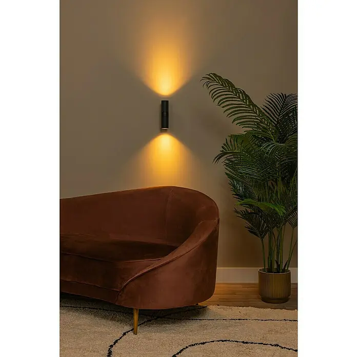 Lucide Clubs Wandleuchte - 50 W, L x B x H: 11,5 x 6,5 x 21,5 cm, Schwarz, Schwarz, GU10 - Wandleuchte in Schwarz, die nach oben und unten gerichtetes Licht spendet, neben einem braunen Samtsofa und einer Zimmerpflanze.