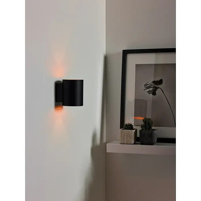 Lucide Wandleuchte Xera - 42 W, L x B x H: 11 x 9 x 11,5 cm, Schwarz, G9 - Wandleuchte aus schwarzem Metall, die ein warmes, orangefarbenes Licht ausstrahlt, neben einem Regal mit Kakteen und einem gerahmten Bild.