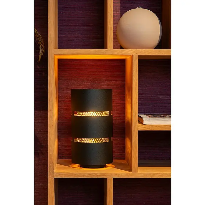 Lucide Rosas Tischleuchte rund - 40 W, Ø x H: 12 x 30 cm, Schwarz/ Gold, Schwarz, E27 - Elegante Tischleuchte mit zylindrischem Design und durchbrochenen Elementen, die ein warmes Licht erzeugen, platziert in einem Holzregal.