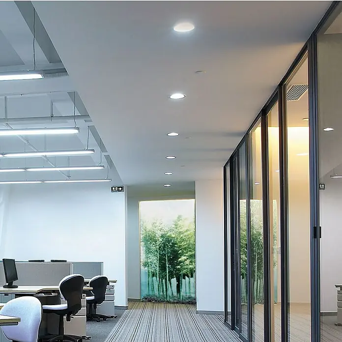 Panaled Downlight empotrable LED redondo Ofis - 6 W, Ø x Al: 12 x 1,8 cm, Blanco cálido - Vista interior de una oficina moderna con luces de techo elegantes, divisiones de vidrio y puestos de trabajo cómodos.