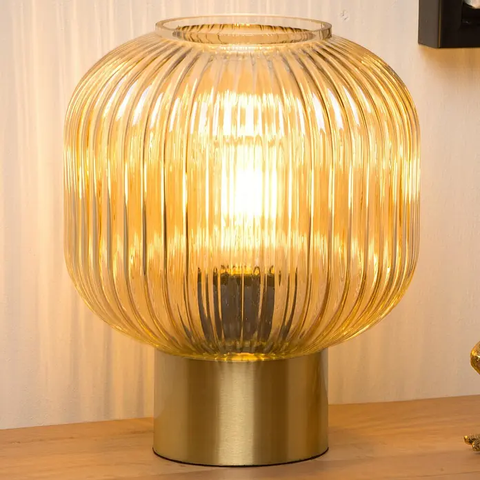 Lucide Maloto Tischleuchte - 40 W, L x B x H: 25 x 25 x 30 cm, Bernstein, E27 - Nahaufnahme einer goldenen Tischleuchte mit einem gerippten Glasschirm und einem goldenen Sockel auf einem Holztisch.