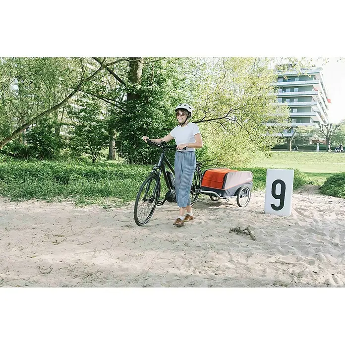 Croozer Fahrradanhänger Cargo Pakko - Traglast: 45 kg, Farbe: Campfire red - Eine Frau mit Helm schiebt ein Fahrrad mit Anhänger auf einem Sandweg. Der Anhänger ist orange und grau.