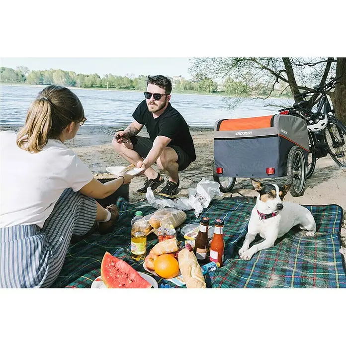 Croozer Fahrradanhänger Cargo Pakko - Traglast: 45 kg, Farbe: Campfire red - Ein Croozer Fahrradanhänger neben einer Picknickdecke am Strand, mit Essen, einem Paar und einem Hund. Ideal für Familienausflüge.