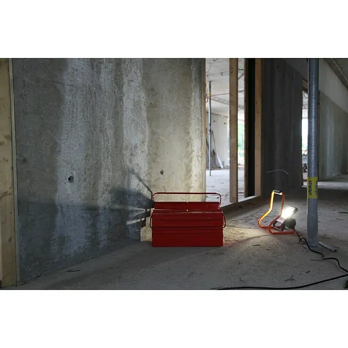 Ledvance LED-Strahler - 30 W, 2.700 lm, 4.000 K, L x B x H: 19 x 22 x 29 cm, IP44 - Roter Werkzeugkasten und eine orangefarbene Arbeitsleuchte auf einer Baustelle. Das Licht beleuchtet den grauen Beton im Raum.