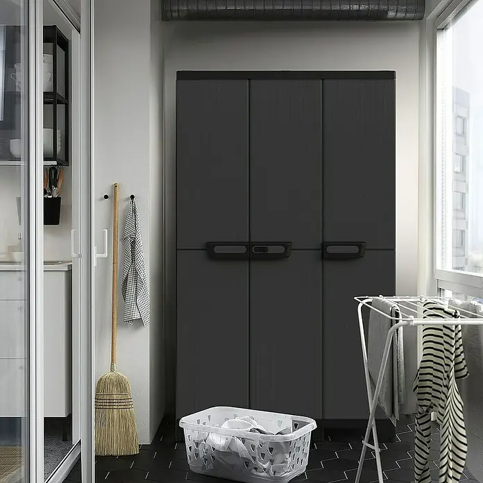 Terry Armario multiusos de plástico  Ardesia - Gris oscuro/Negro, 2 piezas - La imagen muestra un armario de plástico gris de tres puertas sobre un suelo de baldosas oscuras. También se ven una cesta de lavandería y un tendedero.