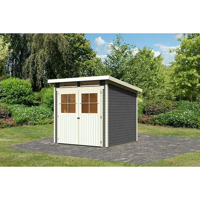 Karibu Gartenhaus Glücksburg - Außenmaß inkl. Dachüberstand (B x T): 242 x 236 cm, Holz, Terragrau, Grundfläche: 4,45 m² - Ein graues Gartenhaus mit weißen Doppeltüren und Fenstern steht auf einem gepflasterten Bereich inmitten eines grünen Gartens.