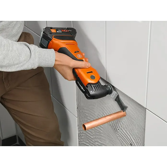 Fein AMPShare 18V Akku-Multifunktionswerkzeug MultiMaster AMM700 MAX AS - 18 V, Ohne Akku, Leerlaufschwingzahl: 10.000 /min - 19.500 /min - Eine Fein Oszillierwerkzeug beim Arbeiten an einer Wand mit einem Kupferrohr. Das Werkzeug ist orange und schwarz.