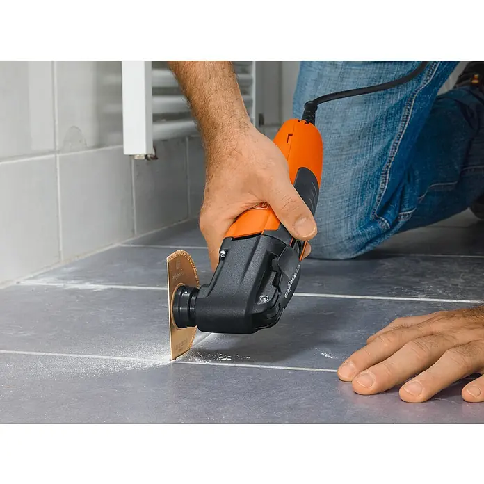 Fein MultiMaster Elektro-Multifunktionswerkzeug MultiMaster MM300 Plus START L-BOXX SET - 250 W, STARLOCK-Plus-System - Multifunktionswerkzeug beim Schneiden von Fugen auf einem gefliesten Boden. Das Werkzeug ist orange und schwarz und hat ein rundes Sägeblatt.