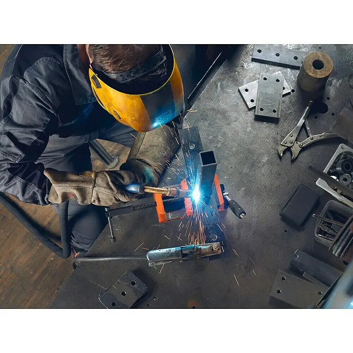 Fein Schraubstock VersaMAG - Spannweite: 108 mm, Metall - Ein Schweißer mit Schutzhelm und Handschuhen bei der Arbeit mit einem Schweißgerät, das helles Licht und Funken erzeugt.