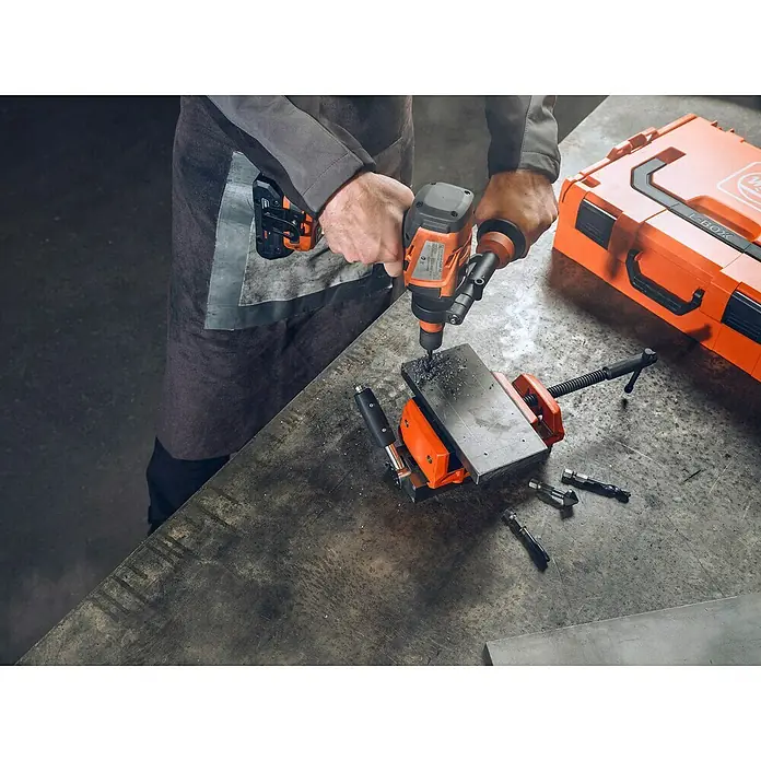 Fein Bohrmaschinenadapter - Passend für: Wechsel von QuickIN Schnittstelle auf die VARIO Schnittstelle, Durchmesser Bohrer: 12 mm - Ein Handwerker bohrt mit einer orangefarbenen Bohrmaschine ein Metallstück, das mit einer Schraubzwinge fixiert ist. Eine Werkzeugkiste steht daneben.
