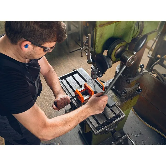 Fein Schraubstock VersaMAG - Spannweite: 108 mm, Metall - Mann an einer Bohrmaschine, der ein Metallstück bearbeitet, Sicherheit geht vor mit Brille und Ohrstöpseln.