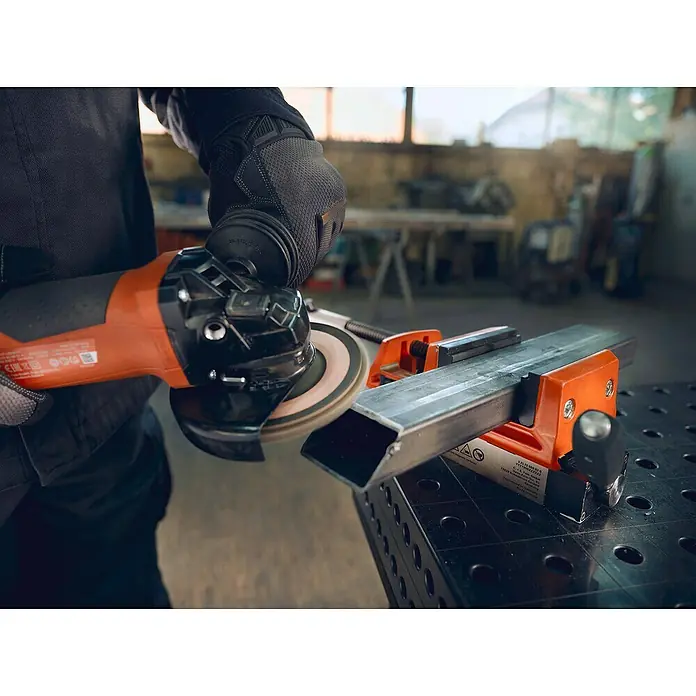 Fein Schraubstock VersaMAG - Spannweite: 108 mm, Metall - Nahaufnahme einer Schleifmaschine, die an einem Metallstück arbeitet, das in einem orangefarbenen Schraubstock befestigt ist.