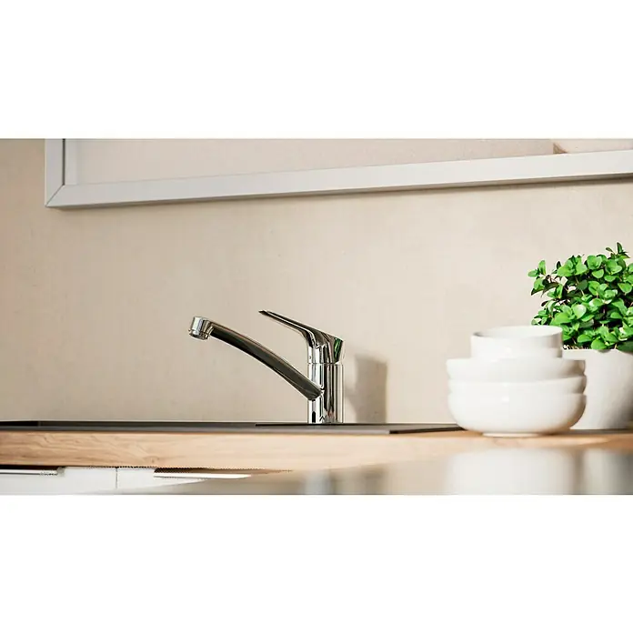 Hansgrohe Grifo de cocina Logis 120 - Cromo, Brillante - Un moderno grifo de cocina cromado combinado con un recipiente de cerámica blanca y una planta verde en una encimera de madera clara.