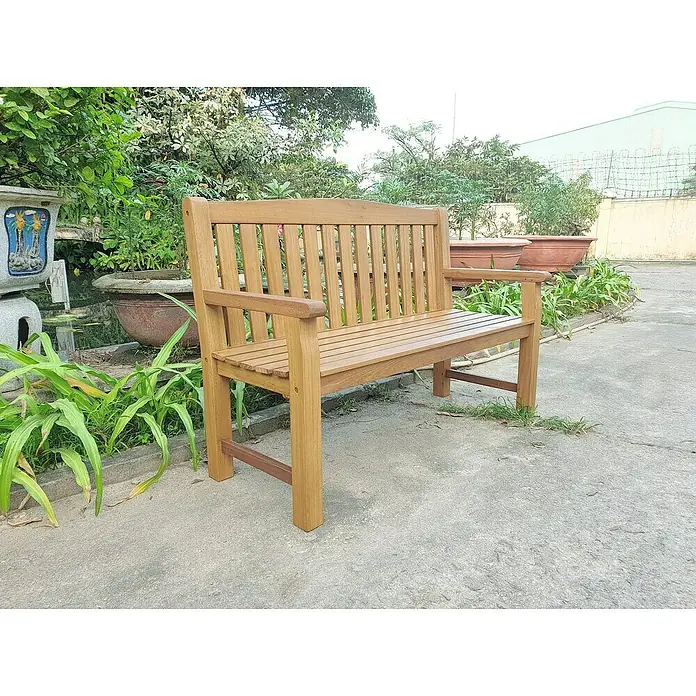 Joda Gartenbank Melbourne 2er-Sitzer - B x T x H: 132 x 58 x 90 cm, Eukalyptus, Hellbraun - Eine hölzerne Gartenbank mit Armlehnen steht auf einem gepflasterten Bereich inmitten üppiger Vegetation. Die Bank lädt zum Verweilen ein.