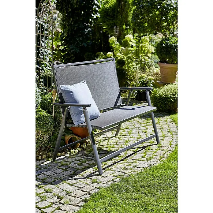 Sunfun Maja Banco de jardín 2 plazas - An x Pr x Al: 119 x 67 x 96 cm, Textilene, Antracita, Abatible - Un banco de jardín gris con un cojín azul en un camino pavimentado en medio de una exuberante vegetación verde.